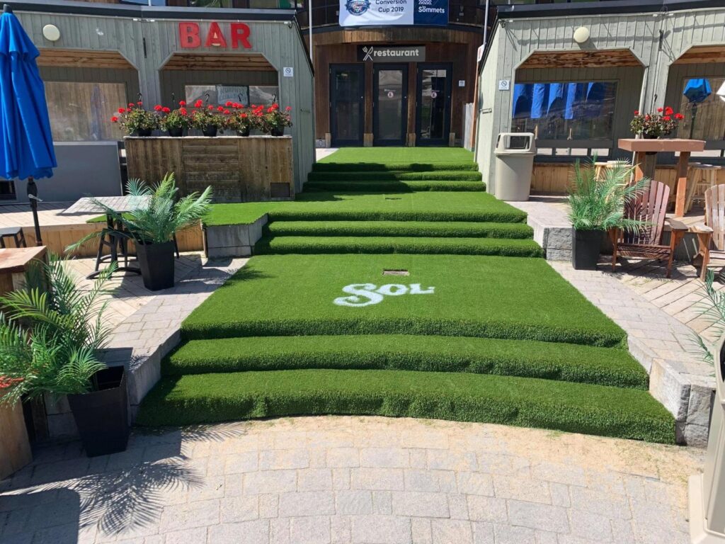 Commercial entrance with artificial grass-covered stairs and pathway, surrounded by outdoor seating, plants, and a restaurant and bar setting.