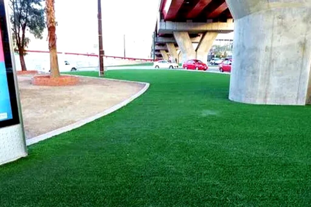 Artificial grass installation under an urban bridge with concrete pillars, creating a clean green landscape in a city environment.
