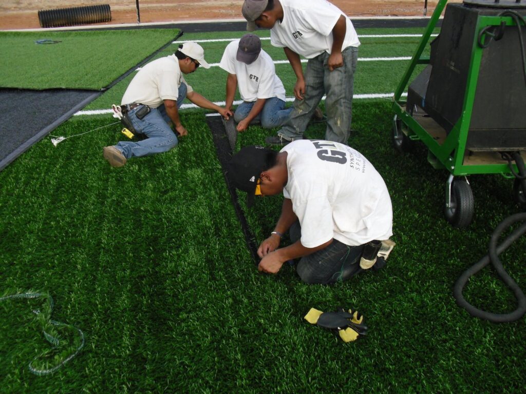Workers installing artificial turf by joining seams on a field, using tools and equipment to secure and align the grass surface.