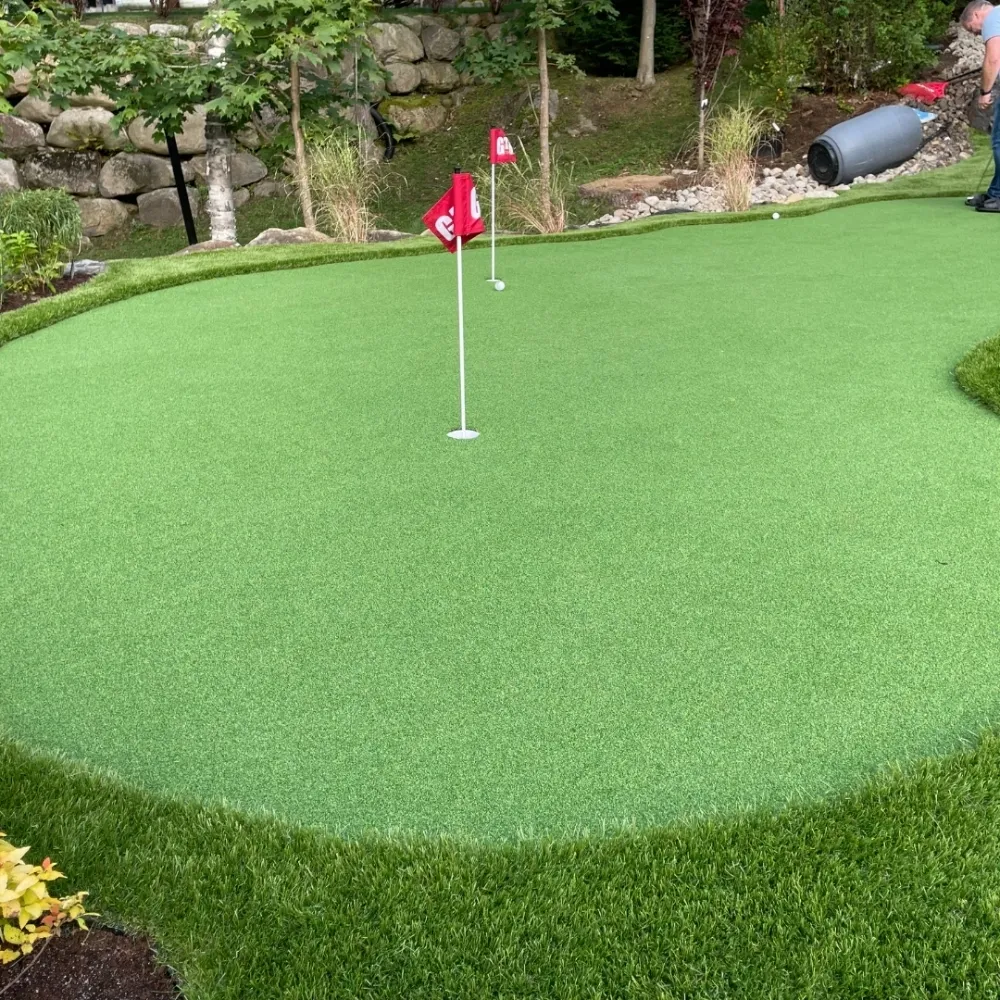 Artificial turf backyard putting green with multiple golf holes and flags, surrounded by landscaping and natural stone elements.