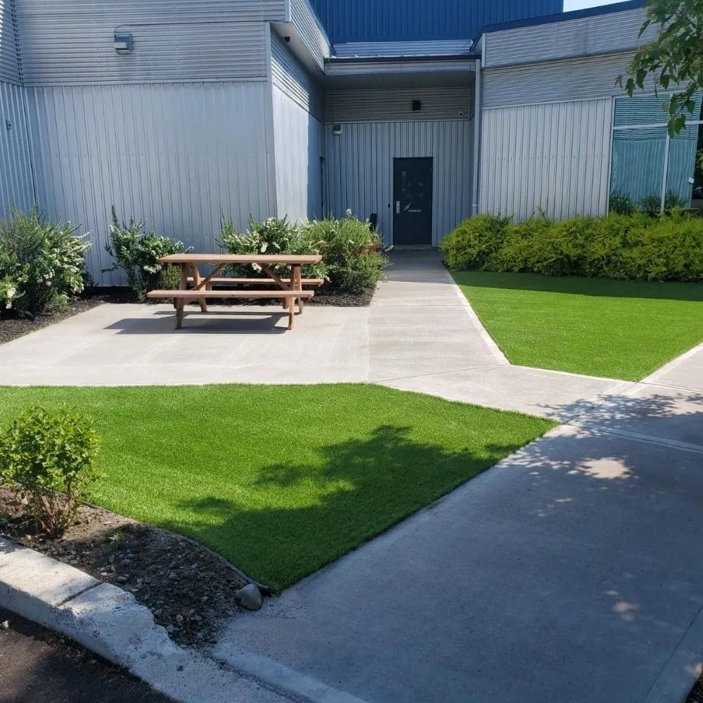 Commercial outdoor area with artificial grass landscaping, concrete pathways, and a picnic table near a building entrance.