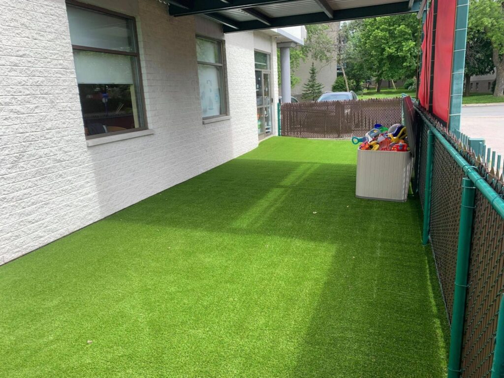Outdoor daycare play area with artificial grass, fenced surroundings, building wall, and toy storage, providing a safe and clean space for children.