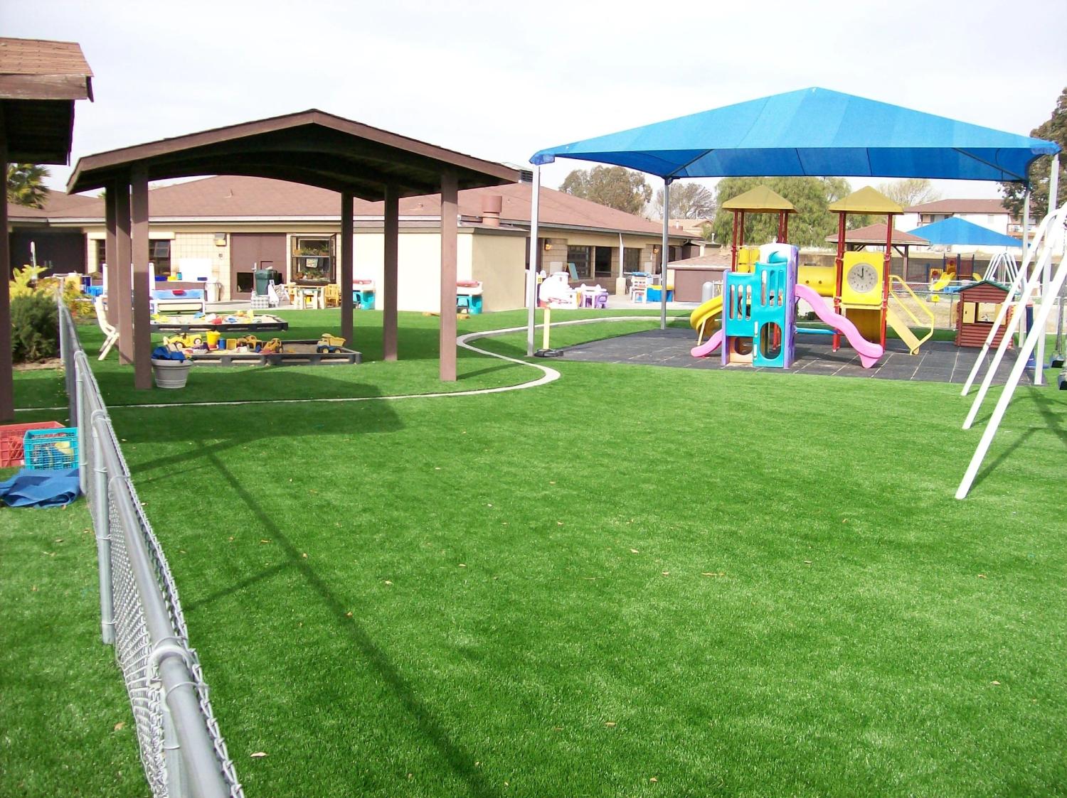 Daycare playground with artificial grass, colorful play structures, shaded seating area, and open outdoor space designed for children’s activities.