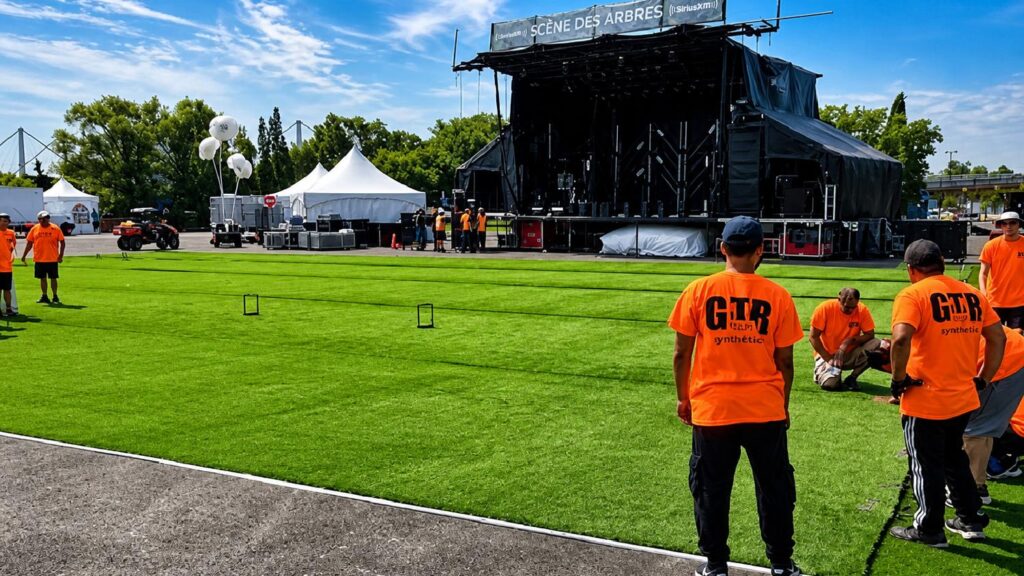 Workers in orange shirts installing artificial turf in front of an outdoor stage labeled “Scène des Arbres,” with tents and event setup in the background.