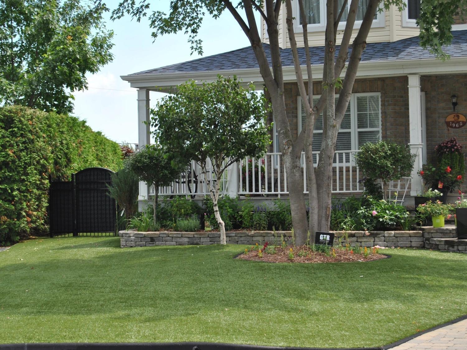 Beautiful residential front yard with artificial grass lawn, mature trees, landscaped garden beds, and suburban home exterior.