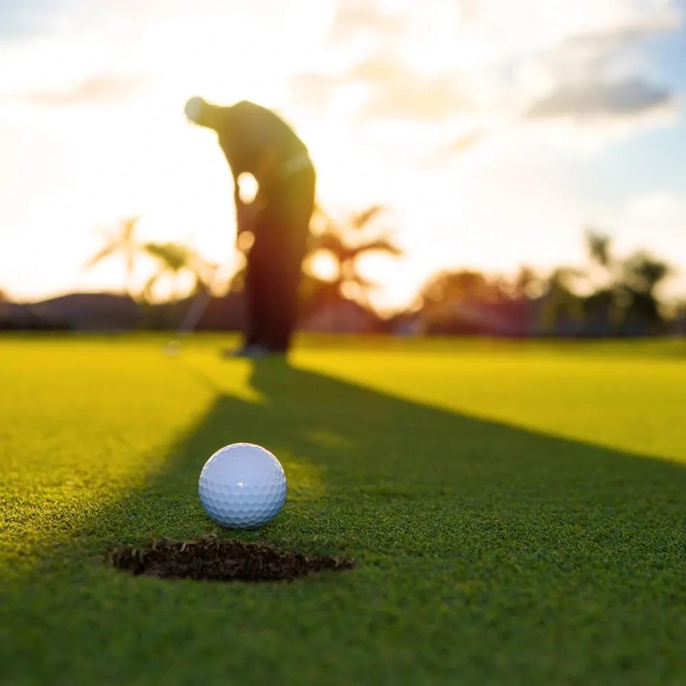 Close-up of a golf ball near the hole on a putting green with a golfer in the background during sunset, highlighting artificial turf performance.