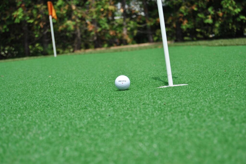 Golf ball on artificial putting green near the hole and flagstick, with smooth turf surface and trees in the background.