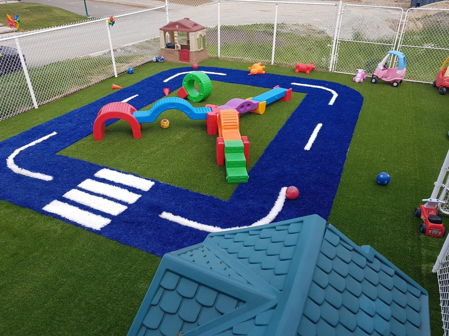 Children’s playground with artificial turf featuring a blue road track design, colorful play equipment, toys, and a fenced outdoor play area.