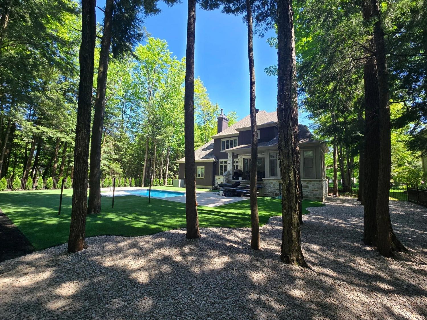 Luxury residential home with wooded backyard, artificial turf lawn, swimming pool, and landscaped outdoor living space surrounded by tall trees.