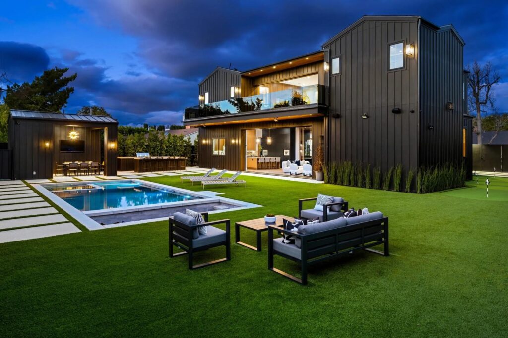 Modern luxury home with artificial grass backyard, illuminated swimming pool, and outdoor seating area at dusk.