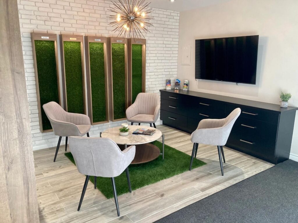 Modern office waiting area with four upholstered chairs around a round table, green decorative wall panels, white brick wall, chandelier lighting, and a wall-mounted TV.