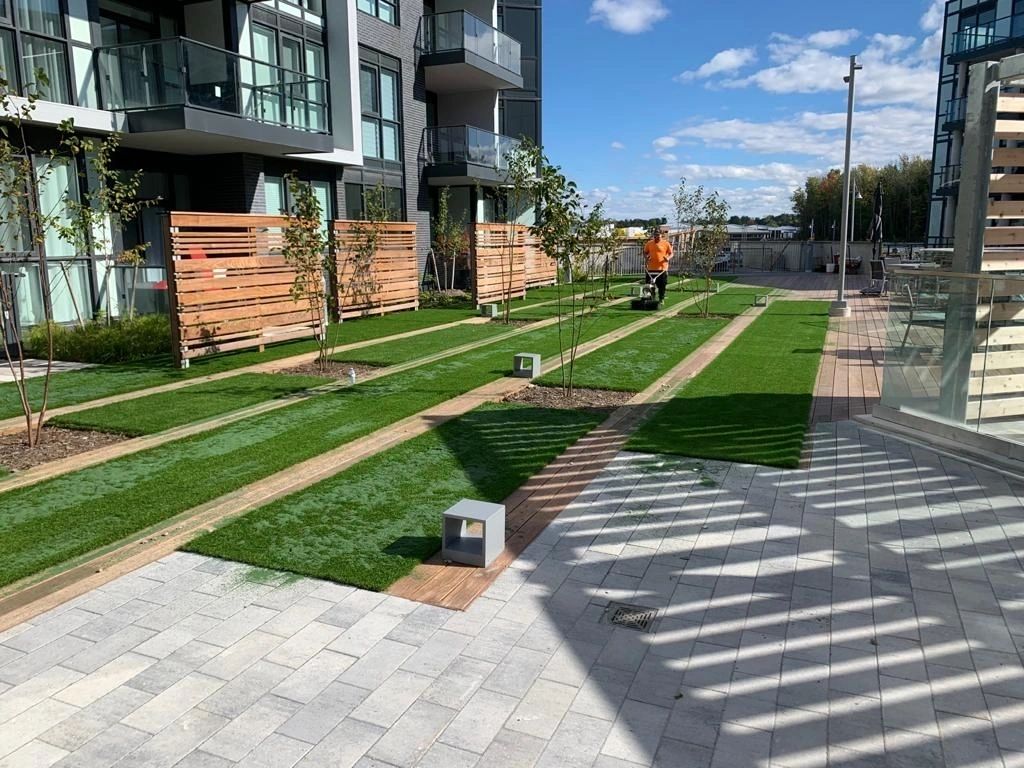 Modern residential outdoor space with artificial grass pathways, landscaped areas, small trees, and apartment buildings with balconies in the background.