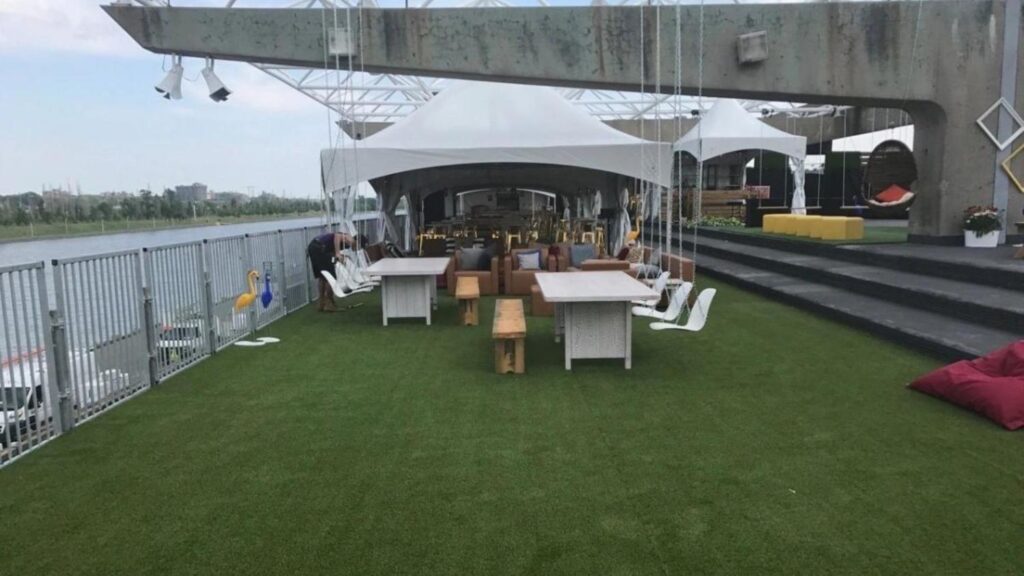 Rooftop event space with artificial turf flooring, white canopy tents, seating arrangements, swings, and waterfront views under an open sky.