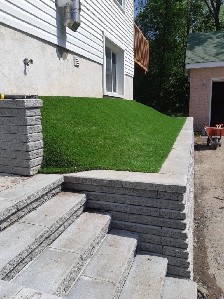 Residential sloped yard with artificial grass installation, retaining wall, stone stairs, and low-maintenance modern landscaping design.