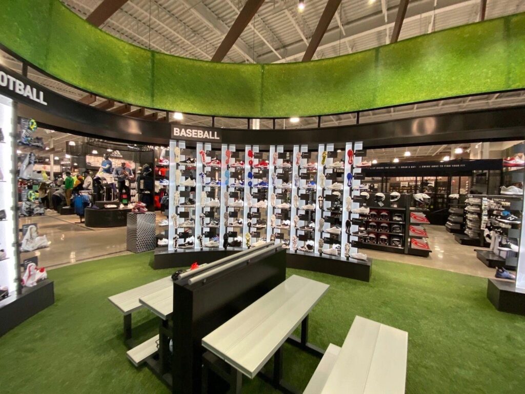Sports retail store interior with artificial turf flooring, shoe display walls, seating benches, and sections for baseball and other sports equipment.