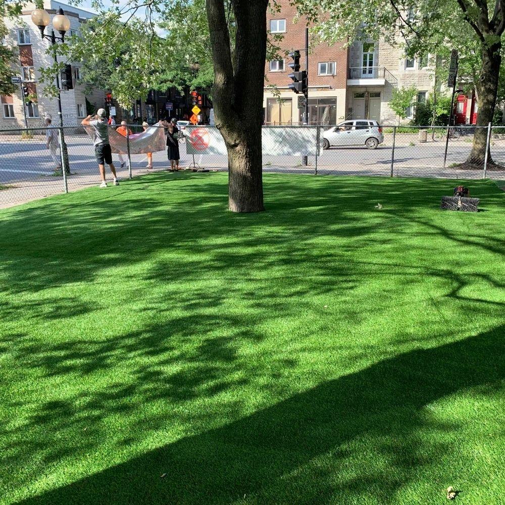 Urban green space with artificial grass lawn around trees, fenced perimeter, and people working in the background near a city street.