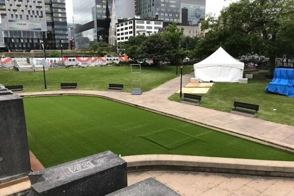 Urban park with artificial grass lawn, curved seating area, pathways, benches, and city buildings in the background with event tents.